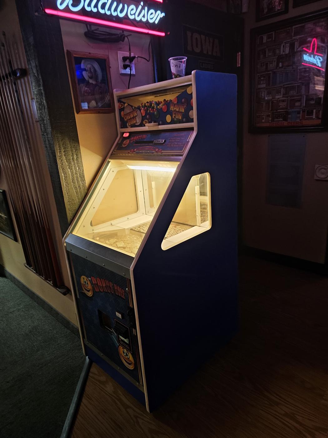 The coin pusher machine beside a rack of pool cues under a Budweiser neon
