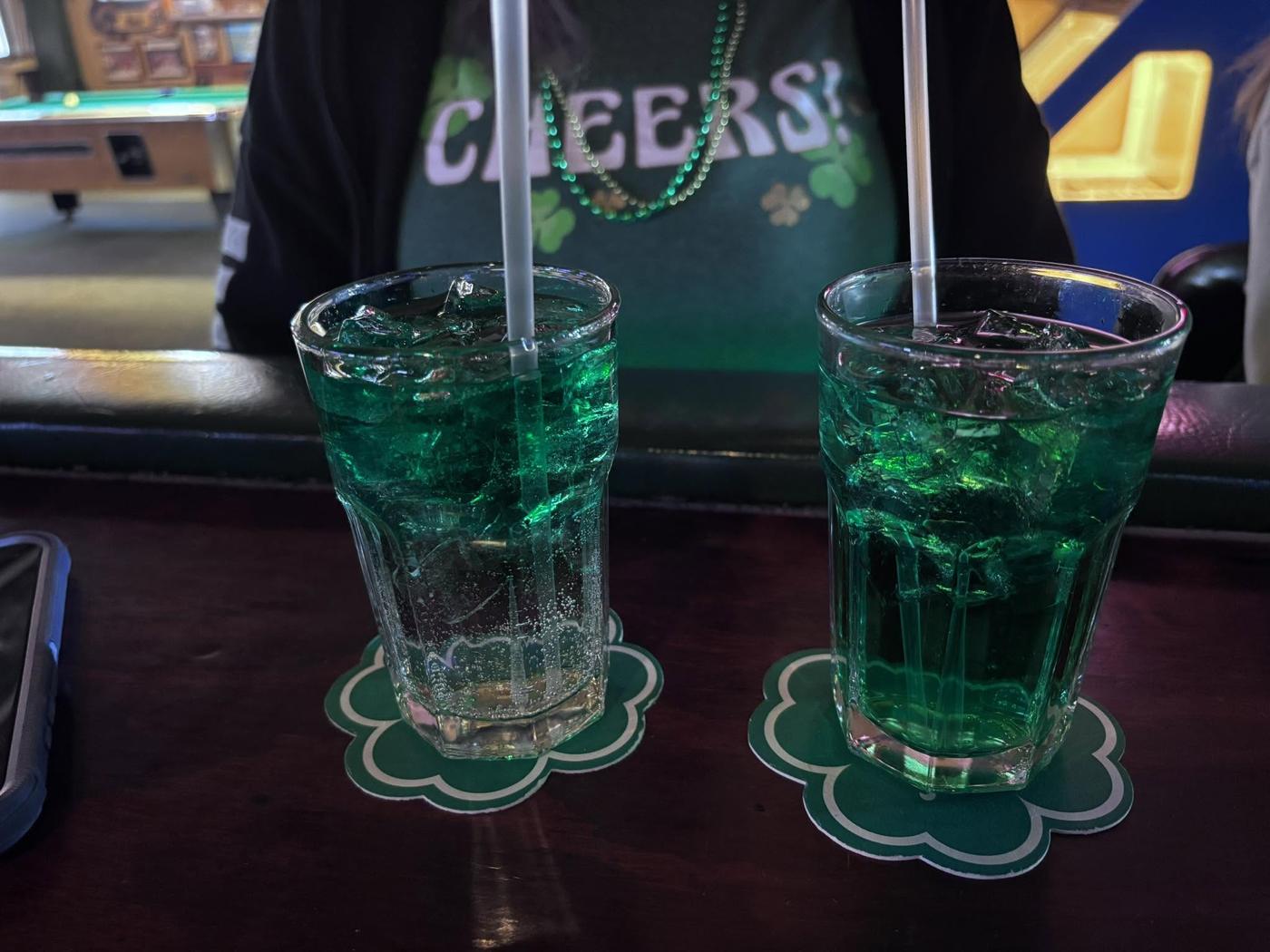 Two green drinks on shamrock coasters with a pool table behind them